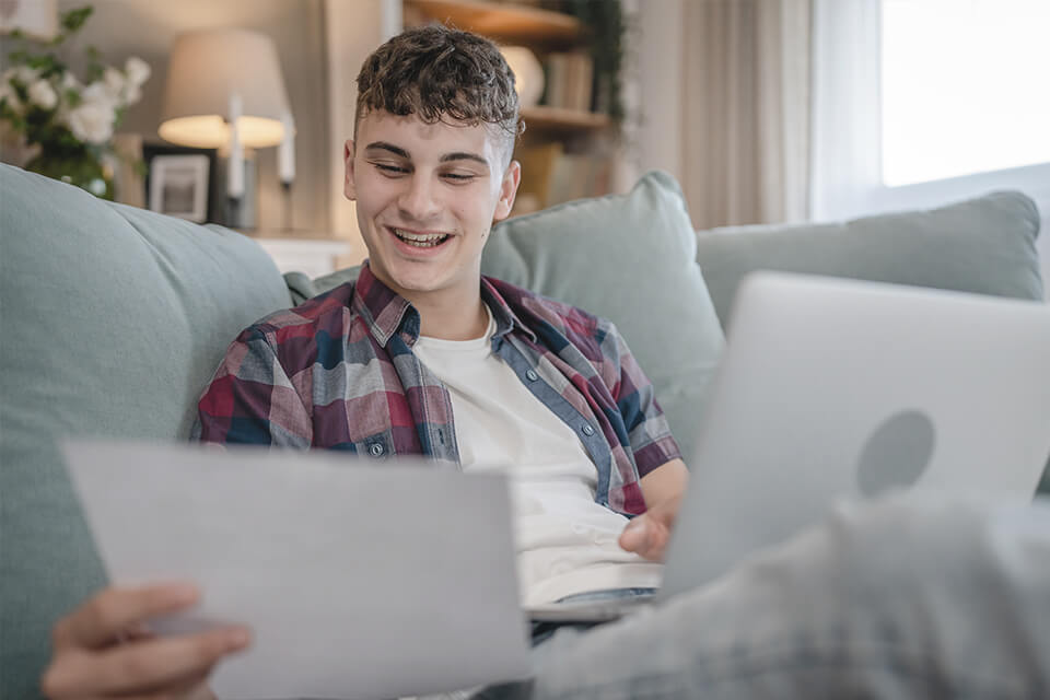 A young man sitting on a couch reviewing a document and a laptop